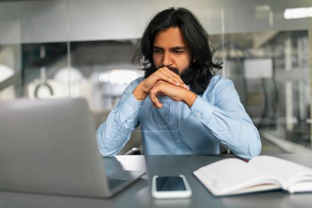 Téléchargez les photos : Homme intensément concentré contemplant son travail sur un ordinateur portable dans un environnement de bureau avec un front sillonné - en image libre de droit