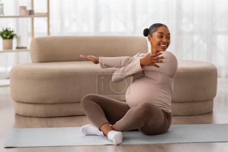 Photo for Pregnant african american woman sitting on yoga mat in living room, stretching arms, copy space. Happy black expecting lady doing sport at home. Healthy lifestyle during pregnancy - Royalty Free Image