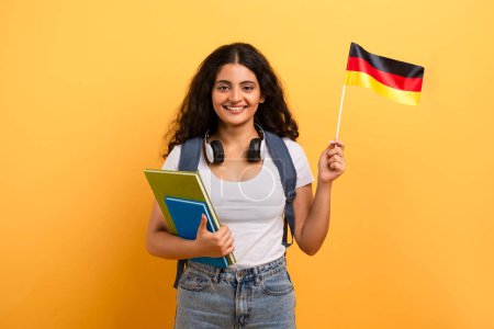 Téléchargez les photos : Une étudiante heureuse avec écouteurs tient un drapeau allemand et des cahiers colorés, suggérant l'apprentissage des langues étrangères - en image libre de droit