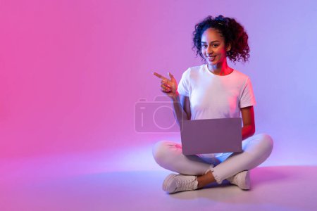 Téléchargez les photos : Joyeux jeune femme noire aux cheveux bouclés, en utilisant un ordinateur portable tout en étant assis jambes croisées et pointant vers le côté à l'espace libre sur fond néon dégradé rose à bleu - en image libre de droit