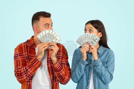 Photo for Excited millennial couple holding dollar money cash, celebrating success, posing with many paper banknotes covering their faces over blue studio background. Young family savings concept - Royalty Free Image