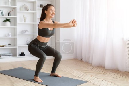 Foto de Mujer asiática enérgica realizando sentadillas ejercicio en casa, joven mujer coreana entrenando en la alfombra de fitness en la sala de estar, sus manos extendidas para el equilibrio, mujer deportiva disfrutando de un estilo de vida activo, espacio de copia - Imagen libre de derechos