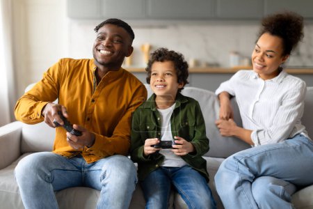 Téléchargez les photos : Une famille est absorbée dans une séance de jeu amusante ensemble sur le canapé, avec des contrôleurs de jeu à la main - en image libre de droit
