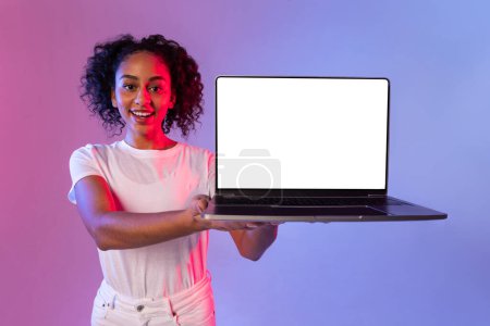 Téléchargez les photos : Joyeux bouclé femme noire offrant un ordinateur portable ouvert avec écran blanc, parfait pour les maquettes, contre dégradé rose et bleu néon toile de fond - en image libre de droit