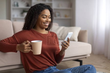 Foto de Ocio doméstico. Mujer negra sonriente relajándose en el piso con teléfono inteligente y café, alegre mujer afroamericana navegando por las redes sociales en el teléfono móvil y disfrutando de la bebida caliente, Copiar el espacio - Imagen libre de derechos