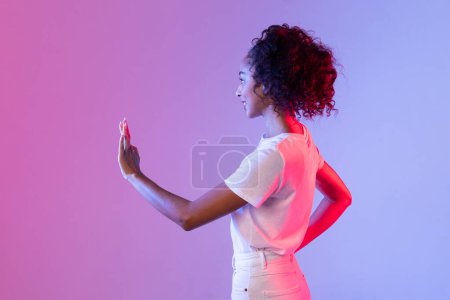 Photo for Young black woman with curly hair focused on an invisible interactive screen, with pink and blue gradient neon background highlighting the scene - Royalty Free Image