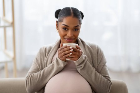 Photo for Portrait of cheerful beautiful young black pregnant woman relaxing on couch at home, drinking tea and smiling at camera. Joyful pregnancy concept - Royalty Free Image