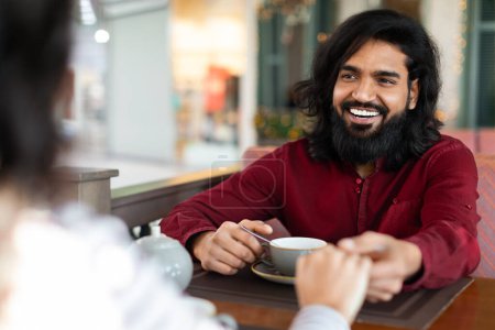 Foto de Alegre barbudo joven indio tiene cita romántica con su novia en el acogedor café, sosteniendo su mano y sonriendo. Millennial pareja disfrutando de tiempo juntos en la cafetería - Imagen libre de derechos