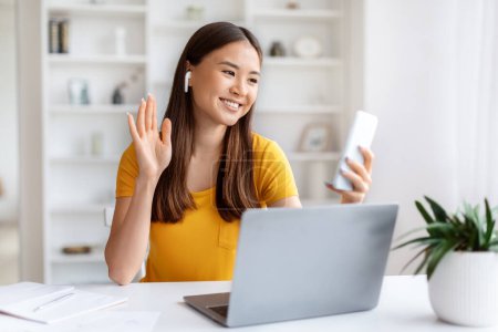 Téléchargez les photos : Délicieuse femme asiatique agitant la main lors d'un appel vidéo sur son smartphone, heureuse jeune coréenne téléconférence sur téléphone portable tout en étant assise au bureau avec ordinateur portable dans son bureau à la maison, espace libre - en image libre de droit