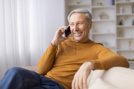 Fröhlicher Senior sitzt auf Couch und unterhält sich angenehm auf seinem Smartphone und reflektiert die Konnektivität