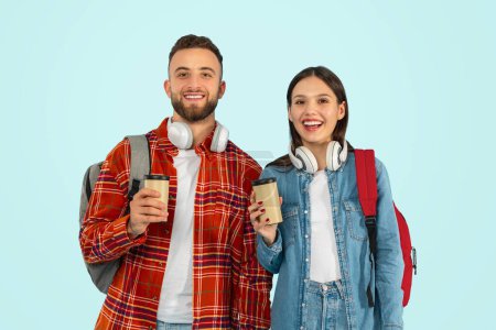 Téléchargez les photos : Couple joyeux d'étudiants posant avec sacs à dos et café à emporter en studio, représentant l'amitié et la convivialité dans leurs années d'université, debout et souriant à la caméra, fond bleu - en image libre de droit