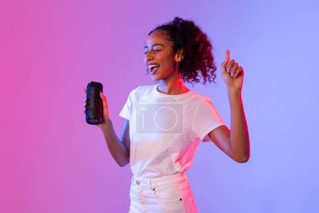 Téléchargez les photos : Jeune femme noire rayonnante aux cheveux bouclés appréciant la musique, dansant tout en tenant haut-parleur portable, sur fond dégradé rose et violet - en image libre de droit