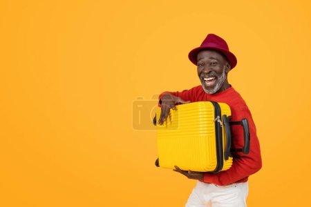 Téléchargez les photos : Joyeux homme noir âgé portant un chapeau rouge et un pull, tenant une valise jaune vif, prêt à voyager avec un sourire joyeux, debout sur un fond orange, studio - en image libre de droit