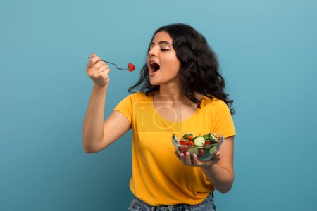 Junge Frau genießt eine Schüssel mit frischem Salat mit fröhlichem Gesichtsausdruck vor blauem Hintergrund