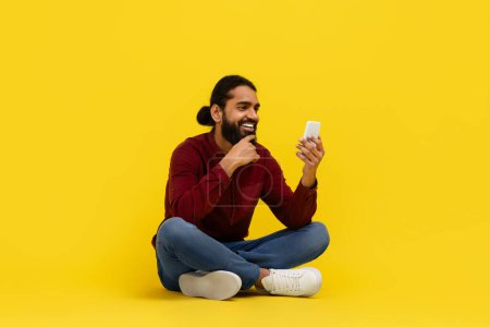Téléchargez les photos : Cool barbu indien gars assis sur le sol isolé sur fond jaune studio, en utilisant smartphone, SMS amis, vérifier la nouvelle application mobile, copier l'espace. Concept de dépendance au gadget - en image libre de droit