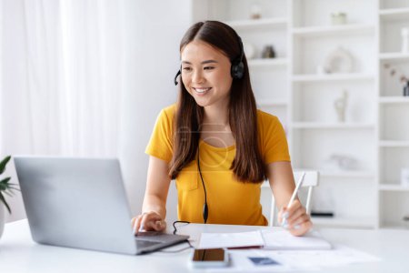 Photo for Joyful young woman wearing headset multitasking with pen and laptop, taking notes while looking at computer screen, korean female working at desk in bright and organized home office, copy space - Royalty Free Image