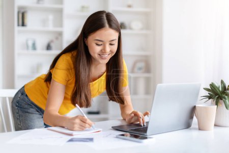 Foto de Mujer asiática sonriente tomando notas mientras trabaja en su computadora portátil en casa, mujer coreana feliz freelancer anotando información de Internet, de pie en el escritorio en la habitación luminosa, espacio de copia - Imagen libre de derechos