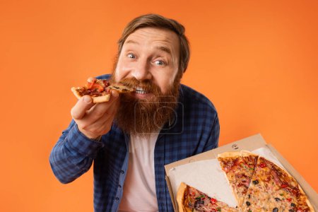 Téléchargez les photos : Barbu Redhaired Guy manger tranche de savoureux Pizza Poser avec boîte délivrée de Pizzeria sur fond de studio orange. Portrait de mangeur appréciant la malbouffe et le repas Cheat. Nutrition, suralimentation - en image libre de droit