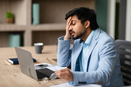 Photo for Workplace Stress. Exhausted Middle Eastern Businessman Touching Face With Tired Expression, Sitting At Laptop During Stressful Work Day In Office, Suffering From Headache And Eyes Fatigue - Royalty Free Image