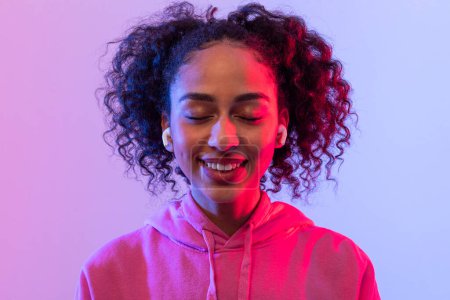 Photo for Relaxed black lady with closed eyes and peaceful smile, wearing earbuds and pink hoodie, against vibrant pink and blue gradient backdrop - Royalty Free Image