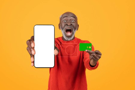 Téléchargez les photos : Étonné heureux surpris homme noir senior présentant un écran vierge smartphone et une carte de crédit, affichant l'excitation et le choc dans un pull rouge sur un fond jaune, studio - en image libre de droit