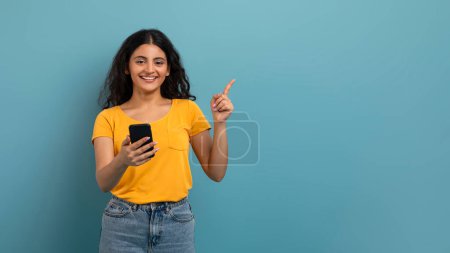 Zufriedene Frau, die ihr Smartphone benutzt und mit einem netten Lächeln auf blauem Hintergrund nach links zeigt