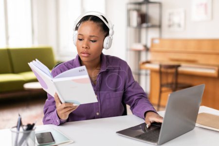 Téléchargez les photos : Étudiante noire concentrée en chemise violette et écouteurs lisant simultanément le livre et l'ordinateur portable, engagée dans le multitâche au bureau à la maison - en image libre de droit