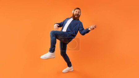 Energetischer Typ mit rotem Bart, der Musik über Kopfhörer genießt, während er unsichtbare Gitarre spielt und sich über orangefarbener Studiokulisse amüsiert. Aufnahme in voller Länge, Panorama mit Kopierraum