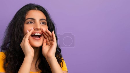 Mujer excitada en una camiseta amarilla grita con las manos tapadas alrededor de la boca, fondo púrpura