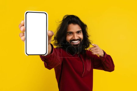 Foto de Cool alegre milenario barbudo indio hombre apuntando al teléfono grande con pantalla blanca en blanco en la mano, recomendando buena aplicación móvil u oferta en línea, fondo amarillo, espacio de copia de maqueta - Imagen libre de derechos