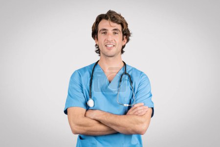 Photo for Smiling male nurse wearing blue scrubs and stethoscope stands with arms crossed, symbolizing professionalism and readiness in medical environment - Royalty Free Image