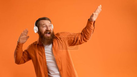 Photo for Party Mood. Cheerful Positive Bearded Guy With Red Hair Listening To Good Music In Wireless Headset, Dancing With Raised Arms And Having Fun Over Orange Studio Wall. Panorama - Royalty Free Image