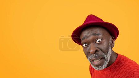 Surpris drôle homme noir senior en chapeau rouge et pull avec un sourcil comique levé, ce qui en fait une expression amusante sur un fond orange solide, studio, panorama, gros plan