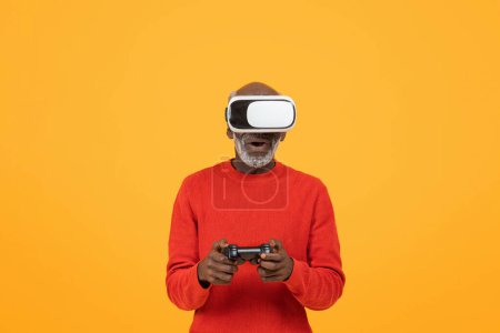 Foto de Sorprendido hombre negro mayor contento usando un auricular de realidad virtual y sosteniendo un controlador de juego, completamente inmerso en una experiencia de realidad virtual interactiva sobre un fondo amarillo, estudio - Imagen libre de derechos