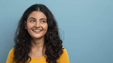 Foto de Una mujer alegre con el pelo largo y rizado con una camiseta amarilla sonríe suavemente sobre un fondo azul sólido - Imagen libre de derechos