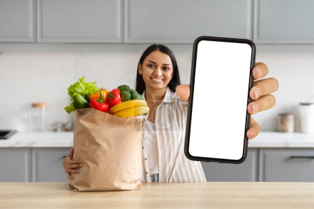Photo for Happy young indian woman order grocery online, showing paper bag full of food and smartphone with white blank screen mockup copy space for delivery mobile app, cozy kitchen interior - Royalty Free Image