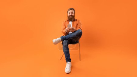 Téléchargez les photos : Redhaired homme sourit tout en textant sur son smartphone dans le studio assis confortablement dans la chaise, communiquer à travers les messages et les médias sociaux, sur fond orange, panorama - en image libre de droit