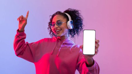Foto de Señora negra vibrante y alegre bailando con auriculares y sosteniendo el teléfono inteligente con pantalla en blanco, contra el fondo dinámico de degradado rosa y azul, que representa el tema de la música y la tecnología - Imagen libre de derechos