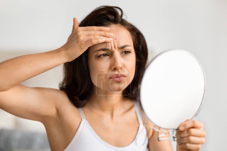 Upset millennial woman looking at mirror, checking pimple on her forehead and grimacing. Unhappy young brunette lady wearing top make daily beauty care, suffering from acne on her face skin