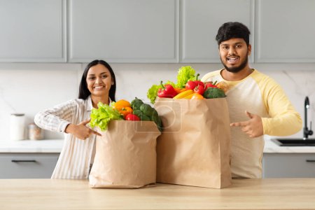 Téléchargez les photos : Positif jeune couple indien pointant vers des sacs en papier d'épicerie pleine de fruits et légumes biologiques frais alimentaires, bénéficiant d'un service de livraison de nourriture à domicile, intérieur de cuisine moderne et confortable - en image libre de droit