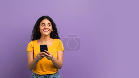 Foto de Mujer joven sonriente usando su teléfono inteligente y mirando hacia arriba con esperanza sobre un fondo violeta - Imagen libre de derechos