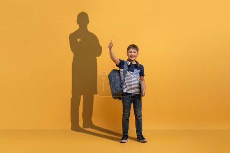 Téléchargez les photos : Un jeune garçon avec un sac à dos donne un pouce devant un fond jaune, avec une ombre suggérant un futur professionnel de la santé, incarnant espoir et ambition. Imagination, avenir - en image libre de droit