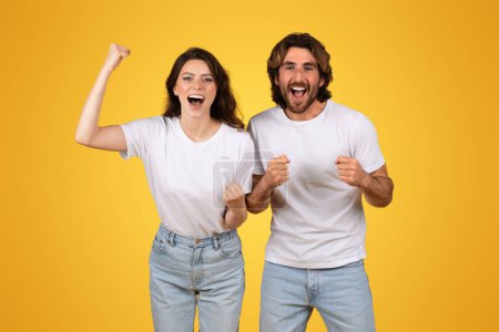 Photo for Energetic european young couple with raised fists celebrating a victory, exuding joy and excitement, wearing white t-shirts and jeans against a vivid yellow background, studio - Royalty Free Image
