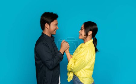 Téléchargez les photos : Un joyeux homme japonais millénaire en chemise sombre et une femme souriante en chemisier jaune se tiennent la main, se regardant avec un sentiment de bonheur et de connexion sur un fond bleu - en image libre de droit