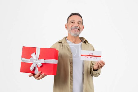 Smiling senior man holding red present box and gift certificate, happy elderly gentleman symbolizing generosity and celebration, standing isolated against white studio background, copy space.