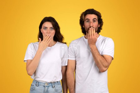 Photo for Shocked millennial european man and a woman with surprised expressions, both covering their mouths with their hands, dressed in white shirts against a yellow background, studio - Royalty Free Image