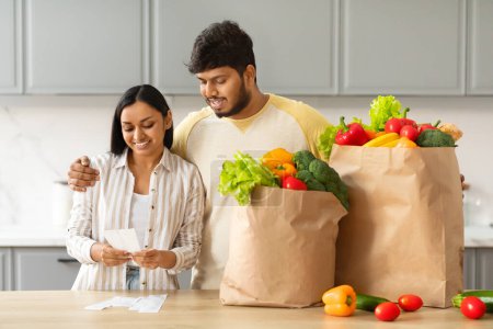 Téléchargez les photos : Heureux jeune homme et femme indien avec des sacs en papier pleins de fruits et légumes biologiques frais alimentaires debout à côté de la table de cuisine, vérifier les factures. Livraison d'épicerie, achats économiques - en image libre de droit