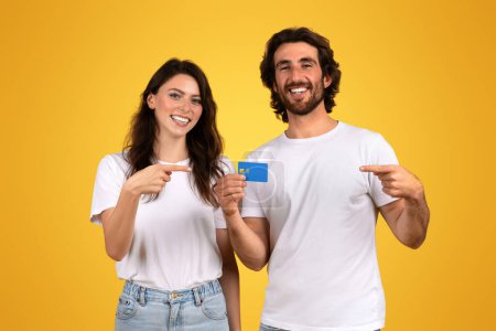 Photo for Smiling young couple in casual white t-shirts, the woman pointing at a blue credit card and the man gesturing towards it, with a bright yellow background symbolizing financial freedom and happiness - Royalty Free Image