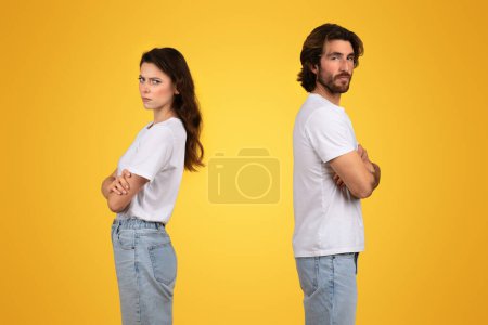 Upset european man and woman standing back-to-back with arms crossed, showing signs of a disagreement or quarrel, wearing casual white t-shirts against a yellow backdrop, studio