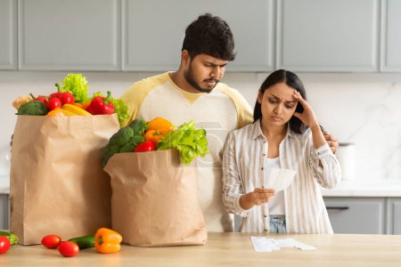 Téléchargez les photos : Frustré malheureux couple indien millénaire avec des sacs en papier plein d'aliments biologiques vérifier les factures après l'épicerie, conjoints choqués par les prix élevés, intérieur de la cuisine confortable - en image libre de droit